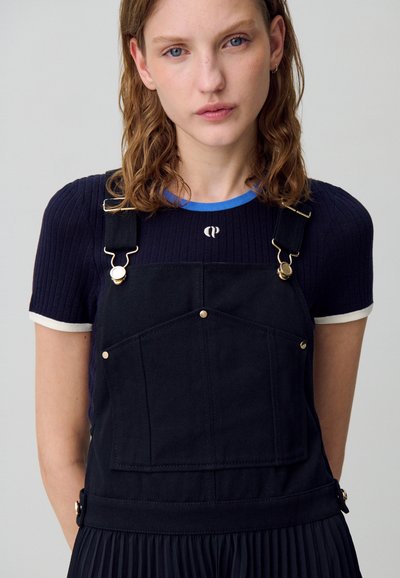 Femme aux cheveux ondulés châtain porte un t-shirt à manches courtes côtelé bleu marine sous des salopettes en denim noir avec des fermoirs dorés et une poche avant.
