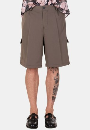 Uomo con indosso pantaloncini cargo marroni, scarpe nere intrecciate e una camicia floreale, che mostra un tatuaggio geometrico su una gamba inferiore.
