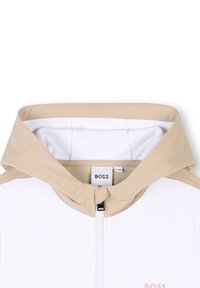 Felpa bianca con zip, dotata di un cappuccio foderato in rete beige e accenti sulle spalle. Logo "BOSS" ricamato sul petto. Texture morbida e liscia.