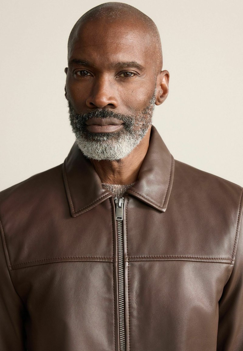 Barna bőrkabát cipzárral, széles gallérral és varrás részletezéssel a mellkason. Simább textúrával és testre szabott fazonnal rendelkezik.