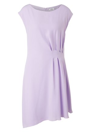 Kobieta ubrana w bezrękawową, lawendową sukienkę do kolan z plisami po bokach, w beżowych plecionych sandałach na obcasie, stojąca z rękami na biodrach.