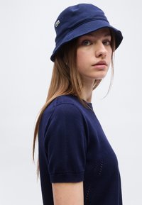 Νεαρή γυναίκα με μακριά μαλλιά, φοράει σκούφο τύπου bucket σε ναυτικό μπλε χρώμα και ταιριαστό κοντομάνικο μπλουζάκι, που κοιτάζει πλευρικά με φόντο έναν απλό τοίχο.