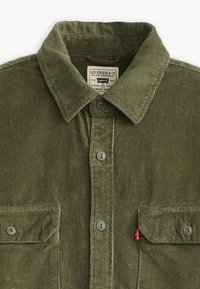 Camicia in corduroy verde oliva con colletto a punta. Presenta due tasche frontali, bottoni grigi e un piccolo etichetta rossa Levi's sulla tasca.