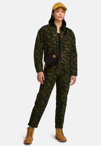 Jumpsuit verde con stampa floreale e zip frontale, abbinato a una borsa a tracolla nera e stivali color cammello. Cappello giallo completa il look.