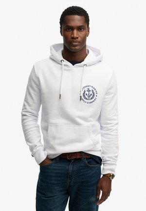 Man die een witte hoodie draagt met een marineblauw ankerlogo, blauwe spijkerbroek, gevlochten riem en polshorloge, staand tegen een effen witte achtergrond.