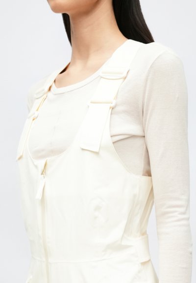 Krém színű nadrágtartós overál állítható pántokkal, cipzáras lezárással és oldalzsebekkel, világos bézs hosszú ujjú felsőre rétegezve. Sima textúrájú.