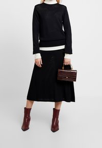 Schwarzer gestrickter Pullover über einem weißen Rollkragenpullover, kombiniert mit einem schwarzen Rock. Burgunderfarbene Ankle Boots und eine strukturierte braune Handtasche runden das Outfit ab.
