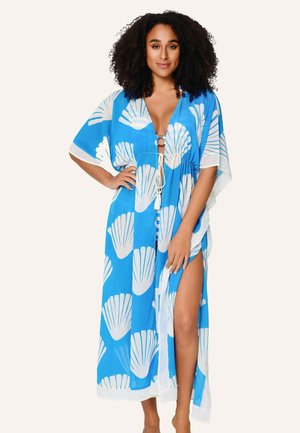 Žena s kučeravými vlasmi, oblečená v modro-bielom plážovom plášti s motívom mušlí, so hlbokým výstrihom do V a bočným rozparkom, stojaca s jednou rukou na boku.