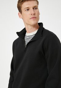 Svart zip-up fleece-tröja med hög krage, tillverkad av mjukt material. Under den syns en vitrandig crew neck-tröja vid halsringningen.