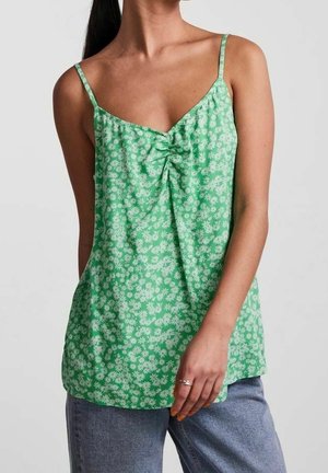 Kobieta ubrana w zielony top bez rękawów z białym kwiatowym wzorem i marszczonym przodem, zestawiony z jasnoniebieskimi dżinsami.