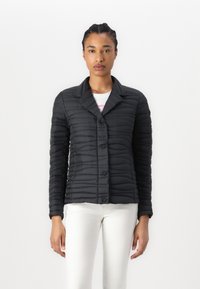 Casaco acolchoado preto com fecho de botões, gola e mangas longas. Apresenta um design texturizado e canelado, sendo combinado com uma blusa branca e calças.