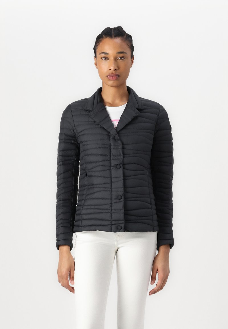 Casaco acolchoado preto com fecho de botões, gola e mangas longas. Apresenta um design texturizado e canelado, sendo combinado com uma blusa branca e calças.