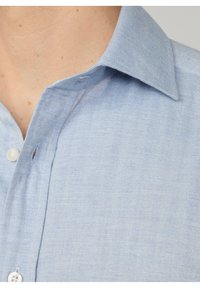 Camisa azul claro, de botones y con un patrón de espiga, que presenta un cuello estándar y botones de plástico. El material parece texturizado y tejido.