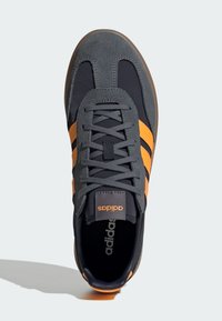 Sneaker with dark blue fabric and grey suede, featuring orange stripes and laces. Includes a black insole with white branding.