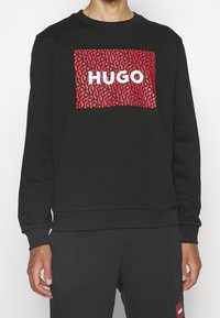 Sudadera negra con un gráfico rojo audaz que repite el texto "HUGO". Cuello y puños de canalé, tela suave, corte relajado.