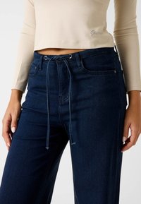 Jean large en denim bleu foncé avec taille à cordon de serrage, comprenant deux poches avant et une texture lisse. Associé à un haut beige à manches longues.