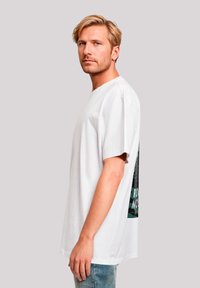 Blond man with blue eyes wearing a white oversized T-shirt and blue jeans, standing sideways against a plain light gray background.