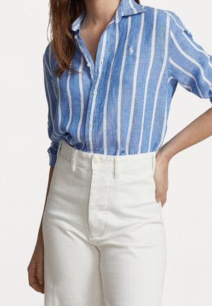 Femme portant une chemise boutonnée à rayures verticales bleues et blanches, rentrée dans un pantalon blanc taille haute, avec une main sur la hanche.