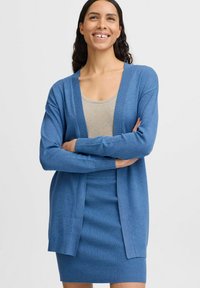 Lichtblauwe cardigan met een open voorkant, lange mouwen en ribbeldetail. Draagt over een beige top en een bijpassende ribbelrok.