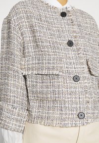 Texturované tweedové sako v béžové barvě s modrými a fialovými vlákny. Má croppovaný střih, třásně na okrajích a černé knoflíky. Obsahuje našité kapsy.