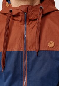 Gros plan sur une personne portant une veste zippée marron et bleu marine avec capuche et cordons, ornée d'un patch logo rond sur la poitrine.