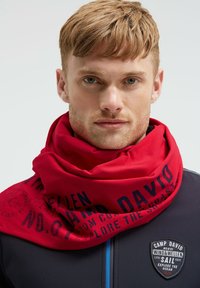 Young man with short hair wearing a red graphic neck scarf and a dark blue jacket with a "Camp David" logo patch.