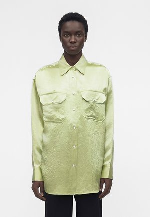 Personne à la peau foncée portant une chemise ample et brillante vert clair boutonnée avec de grandes poches à rabat et un pantalon noir, tournée vers l'avant sur un fond blanc.