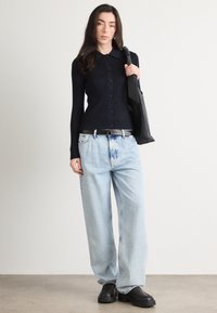 Žena v čiernom zapínacom svetri, svetlomodrých širokých nohaviciach, s čiernym opaskom a čiernymi masívnymi topánkami, nesúca čiernu kabelku na plece.