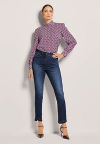 Paarse blouse met een patroon en hoge hals, gecombineerd met donker gewassen skinny jeans en bordeauxrode hoge hakken. Model poseert met één hand op de heup.
