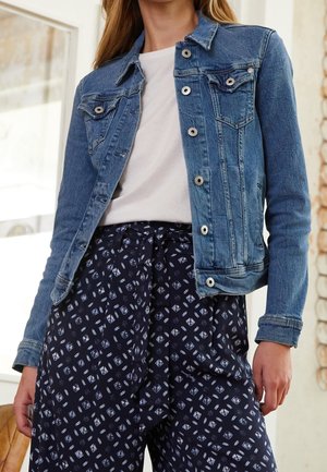 Frau trägt eine blaue Jeansjacke über einem weißen Hemd, kombiniert mit dunklen, gemusterten, hoch taillierten Hosen, die an der Taille mit einem Stoffgürtel gebunden sind.