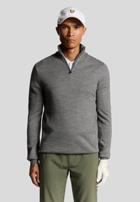 Pullover grigio con mezza zip, realizzato in maglia testurizzata. Abbinato a pantaloni verdi e un cappellino bianco con un logo giallo. Guanto da golf sulla mano sinistra.