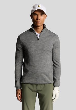 Grau pullover mit halbem Reißverschluss, aus strukturiertem Strick. Kombiniert mit grünen Hosen und einer weißen Mütze mit gelbem Logo. Golfhandschuh an der linken Hand.