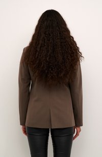 Blazer marrone aderente con una sola fascetta sul retro, abbinato a pantaloni di pelle nera. I capelli sono ricci e scendono oltre le spalle.