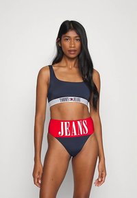 Top bikini blu navy con una banda grigia a contrasto con la scritta "TOMMY JEANS"; slip a vita alta in blu navy e rosso con la grande scritta bianca "JEANS".