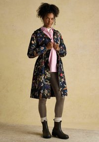 Tânără în picioare, purtând o jachetă lungă de ploaie cu imprimeu floral peste un tricou roz, colanți în carouri, șosete albe și cizme scurte închise la culoare.