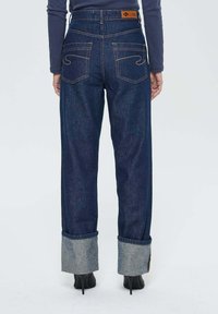 Personne portant un jean bleu marine retroussé et des chaussures à talons noirs, se tenant de dos sur un fond blanc.