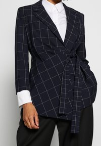 Marinblå kavaj med vita rutiga linjer, med en omslagdesign, långa ärmar och ett knytband framöver en vit skjorta.