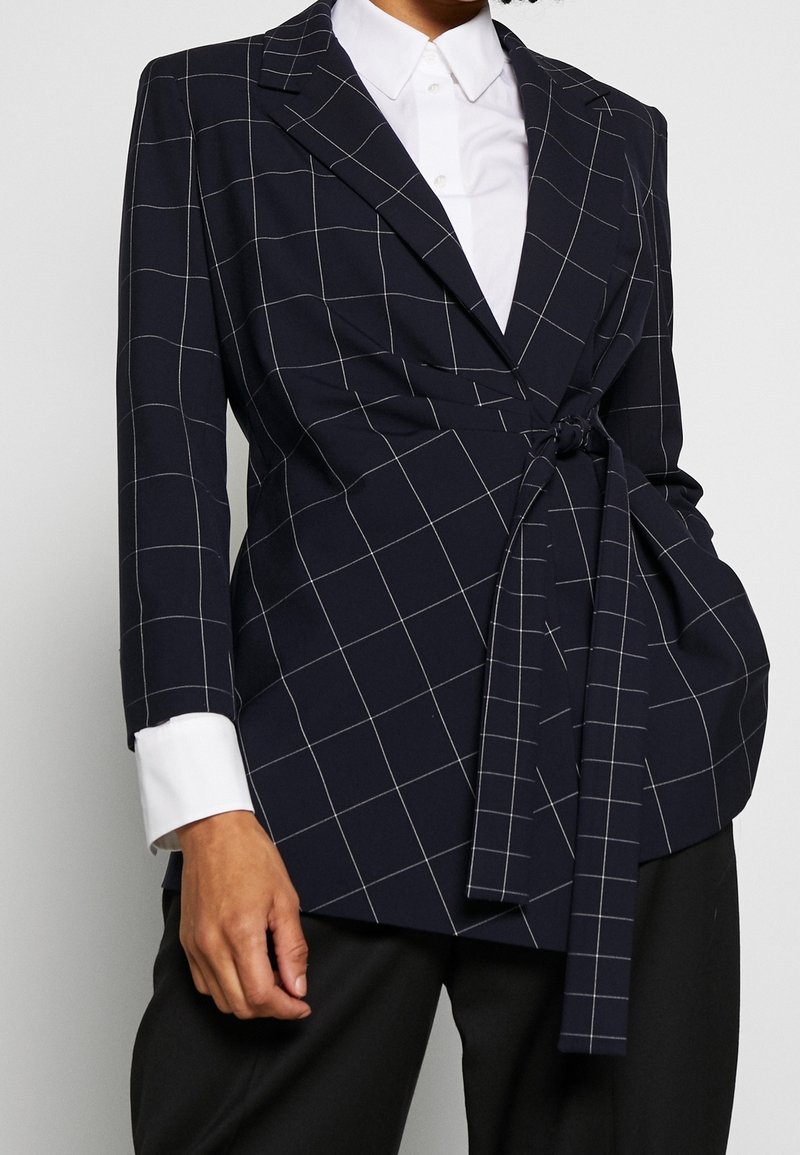 Marinblå kavaj med vita rutiga linjer, med en omslagdesign, långa ärmar och ett knytband framöver en vit skjorta.