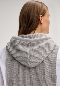 Graue gestrickte Weste mit großem Kapuzen, mit einer gerippten Textur. Das Rückendesign ist nahtlos und liegt über einem weißen Hemd.