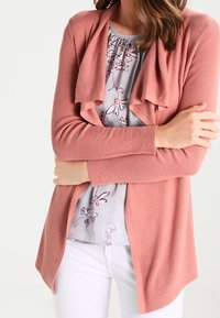 Cárdigan de punto acanalado color rosa con apertura frontal, mangas largas y cuello caído, combinado con una blusa ligera de flores y pantalones blancos.