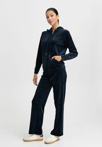 Marinblå velour joggingdräkt med en zip-up huvtröja, framfickor och avslappnade byxor. Mjuk textur och justerbar snörning vid huvan.