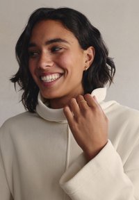 Maglione bianco a coste con collo alto e maniche ampie. La persona indossa piccoli orecchini a cerchio dorati e un semplice anello d'oro sulla mano.