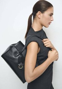 Sac cabas en cuir noir avec une forme rectangulaire, une finition texturée, des anses arrondies et un accent en anneau métallique. Porté à l'épaule.