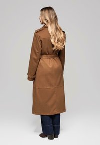 Abrigo largo marrón con cinturón en la cintura, hombreras y una costura en la parte trasera, combinado con pantalones vaqueros de pierna ancha y botas oscuras.