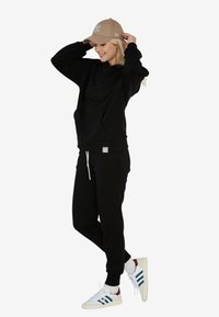 Czarna bluza z kapturem z przednią kieszenią, pasujące czarne dresy i beżowa czapka. Białe sneakersy z niebieskimi i bordowymi paskami, luźny krój.