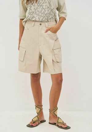 Bézs színű cargo rövidnadrág elöl gombbal, oldalsó hajtókás zsebekkel, grafikus pólóval és arany szegecses pántos szandállal viselve modellen.