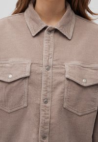 Camicia in corduroy di un leggero taupe con righe verticali, colletto a punta, bottoni a scatto in metallo e due tasche sul petto con patta.