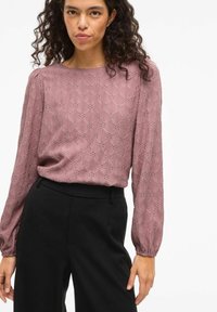 Sieviete ar cirtainiem matiem, tērpusies mauve krāsas mežģīņu raksta garām piedurknēm blūzē un augsto vidukļa melnā biksēs uz viendabīgas baltas fona.