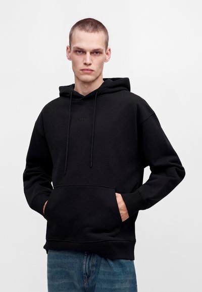 Jeune homme aux cheveux courts portant un sweat à capuche noir et un jean bleu, debout les mains dans les poches devant un fond uni.