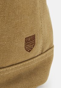 Tejido de lona marrón con un patrón texturizado y tejido, con un distintivo logo bordado de un escudo en marrón más oscuro en la esquina inferior.